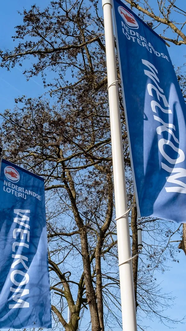 Vlaggen van de Nederlandse Loterij met de tekst 'Welkom' in een blauwe achtergrond en bomen op de achtergrond.