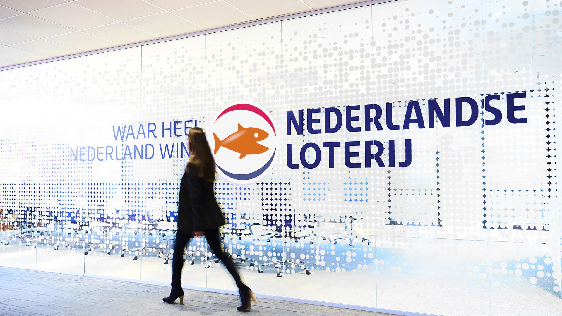 Een vrouw loopt langs een glazen wand met het logo en de naam van de Nederlandse Loterij.