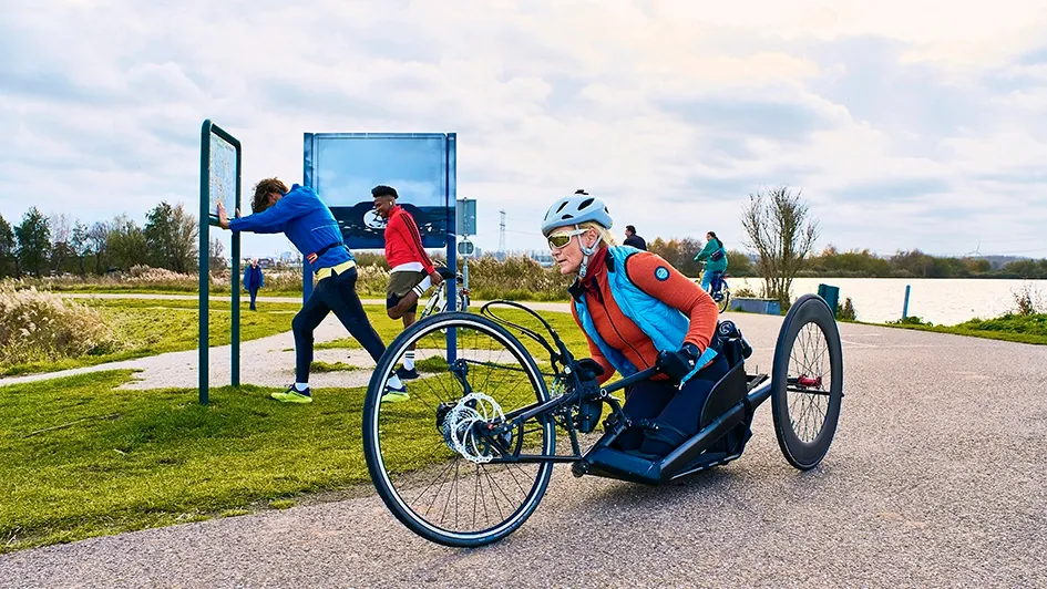 Een persoon in een handbike, terwijl anderen op de achtergrond wandelen en een informatiebord bekijken.