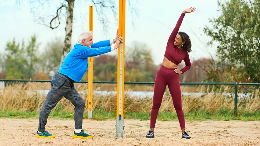 Twee mensen die oefeningen doen op een buitensportlocatie, de een rekt zich uit en de ander steunt op een paal.